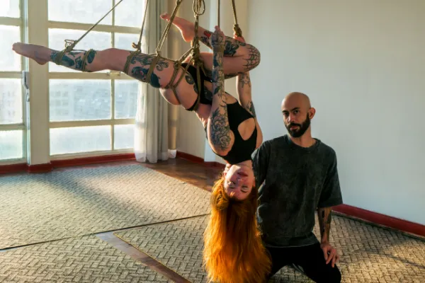 Mulher tatuada suspensa em shibari com cordas de juta, de cabeça para baixo, ao lado de um homem, em ambiente iluminado por janela.