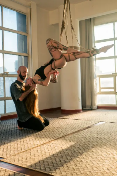 Homem ajoelhado sustenta mulher suspensa em shibari, amarrada com cordas de juta. Ambiente iluminado com janelas amplas, luz natural e estética artística.