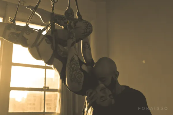 Pessoa tatuada suspensa em shibari com cordas de juta. Homem a beija em ambiente iluminado pela luz de uma janela, estilo sépia.