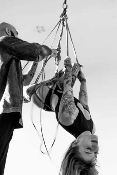 Praticante de shibari ajusta cordas de juta em pessoa tatuada suspensa, em pose artística, com iluminação clara, estilo monocromático.
