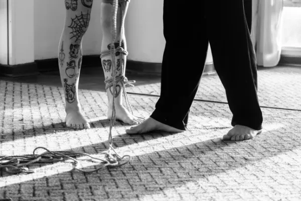 Pernas tatuadas em sessão de shibari com cordas de juta amarradas, ao lado de pessoa descalça em ambiente iluminado. Registro em preto e branco.