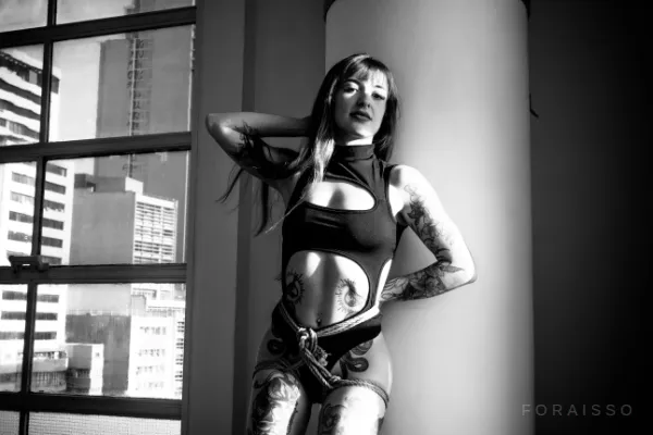 Mulher tatuada posando em ambiente urbano com cordas de juta amarradas ao corpo, estilo shibari, em iluminação contrastante de preto e branco.