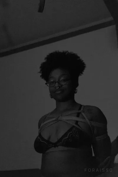 Pessoa de óculos com cabelo crespo, vestindo lingerie de renda e amarrações de shibari com cordas de juta sobre o busto em ambiente de baixa luz.