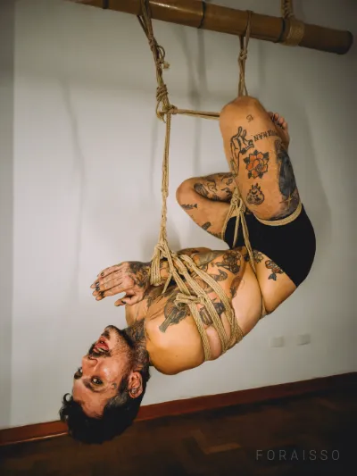 Homem tatuado em suspensão shibari, suspenso por cordas de juta amarradas a um bambu, em fundo neutro com iluminação suave.