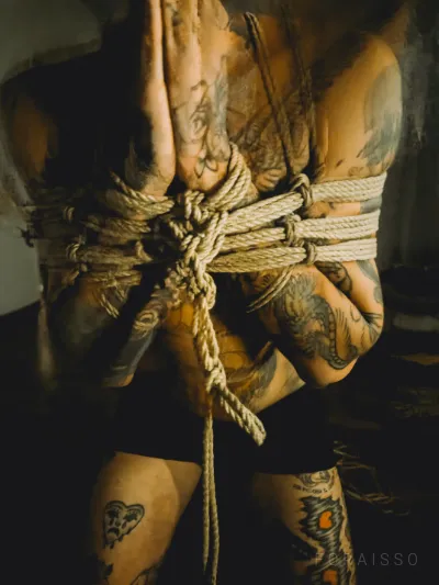 Pessoa tatuada em shibari com cordas de juta. Tronco e braços amarrados, mãos unidas. Luz dramática.