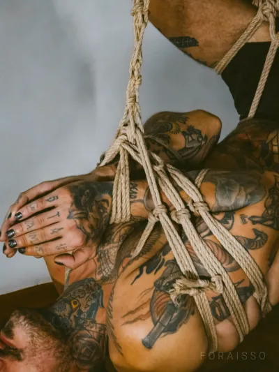 Pessoa tatuada suspensa em shibari elaborado de cordas de juta. Corpo amarrado e invertido, unhas escuras.