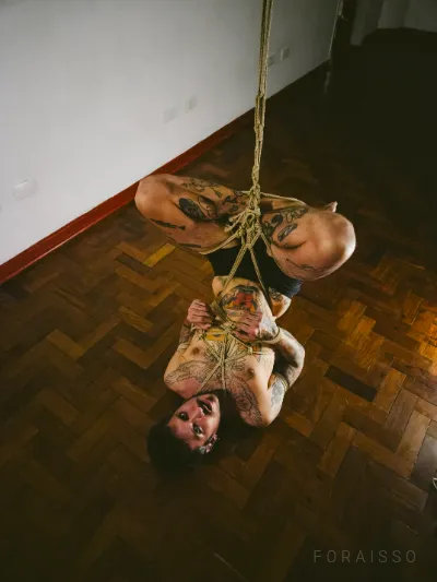 Homem tatuado suspenso em shibari de cabeça para baixo, amarrado com cordas de juta sobre chão de madeira.