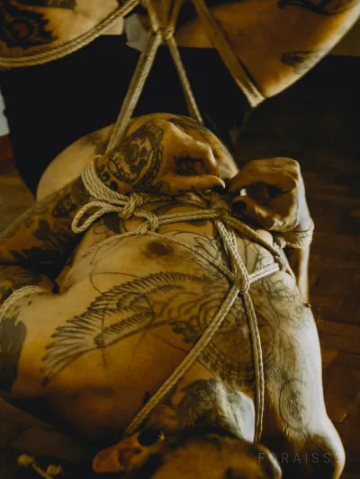 Pessoa tatuada em shibari com cordas de juta. Mãos atadas ao corpo, iluminação quente e íntima.