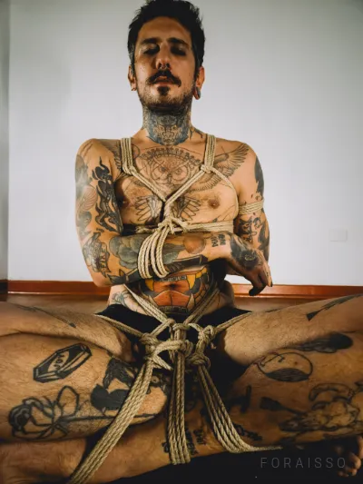 Homem tatuado em shibari, amarrado com cordas de juta no tronco e pernas, sentado. Fundo claro.